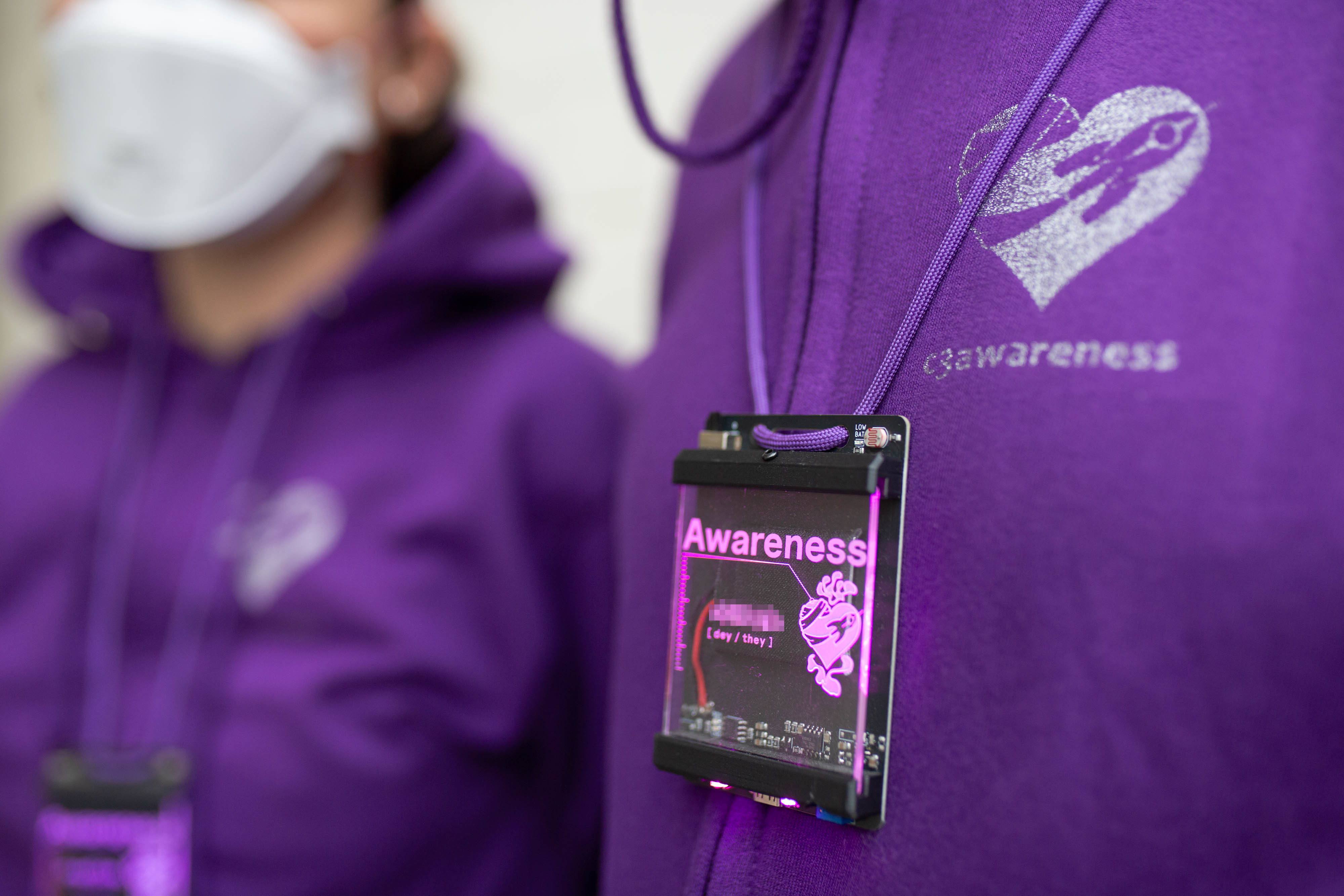 The photograph shows the torso of two individuals wearing purple hoodies and electronic badges which light up purple. On both hoodies and badges the c3awareness logo is visible: a fairy dust rocket is bandaging a heart. On the badge the title 'Awareness' is lit up and below a pixelated name and pronouns are visible. Photo (c) c3awareness | TRim