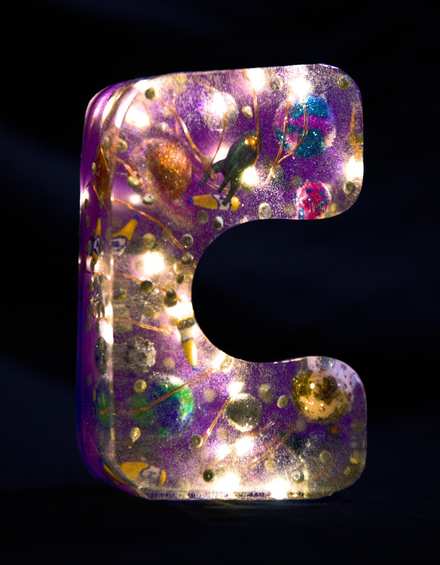 A 'C' moulded from epoxy resin. Inside, a string of lights glows and small planets, mate bottles, a rocket and glitter particles float around. The letter is dark purple in colour. Foto (c) c3awareness | TRim