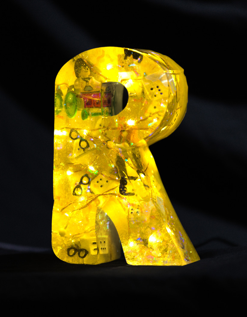 An 'R' made of epoxy resin. Inside, fairy lights glow, there's a Lego figure in a wheelchair, white Braille keyboard keycaps, and a Lego figure with a white cane and some sunglasses float around inside. The letter is coloured yellow.
        Photo (c) c3awareness | TRim