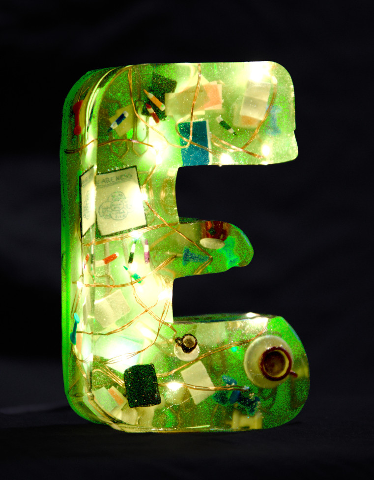 An 'E' made of epoxy resin. Inside, a fairy lights glow, and coffee and tea cups, books, pens, presentation cards, loudspeakers and a whiteboard with a diagram float inside. The letter is coloured light green.
        Photo (c) c3awareness | TRim