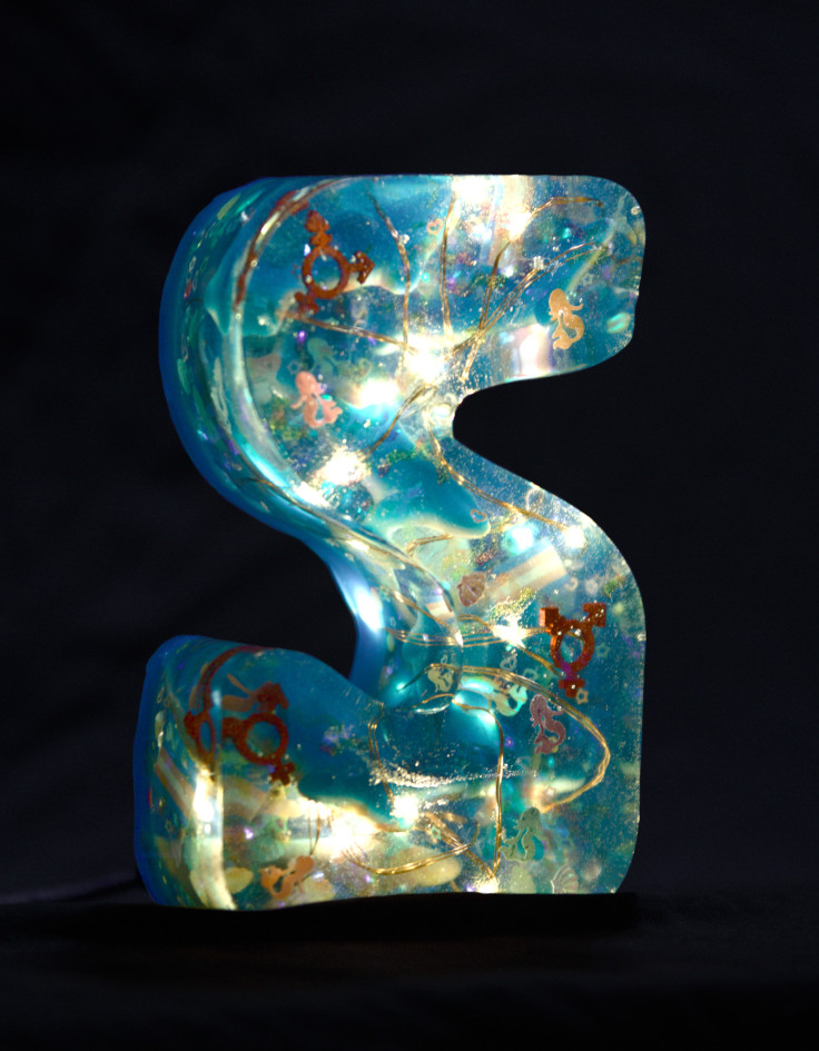 An S cast from epoxy resin. Inside, some fairy lights glow and sharks, shimmering mermaids, small trans pride flags and transgender symbols float around. The letter is light blue in colour.
        Photo (c) c3awareness | TRim