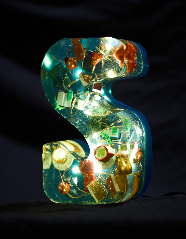 An 'S' cast from epoxy resin. Inside, some fairy lights glow and small teacups, armchairs, boxing gloves, books with bookworms, hot water bottles, fruit and ladybirds float around. The letter is dark blue in colour.
        Photo (c) c3awareness | TRim